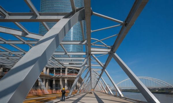 A person in a yellow hard hat stands on a bridge under construction, with steel beams forming a geometric pattern. A modern glass skyscraper and an arched bridge are visible in the background under a clear blue sky.